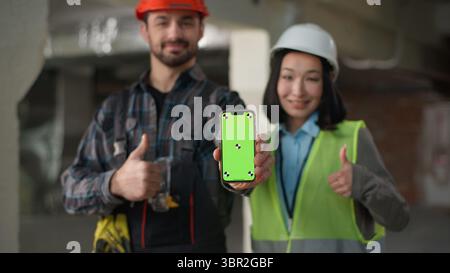Two multiethnic builders using mobile phone building design app showing thumb up Japanese woman Caucasian man hardhat engineers business inspector Stock Photo