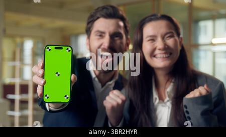 Excited two business woman man celebrating win showing mobile phone green screen chroma key mock up Caucasian Korean businesspeople cheering winner Stock Photo