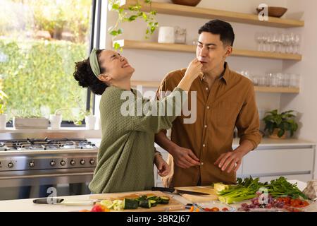 Diverse couple feeding each other chopped zucchini and cherry tomatoes at kitchen island Stock Photo