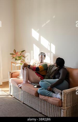 Diverse female friends sitting on sofa at home checking smartphone with potted plant on side table Stock Photo