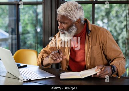 Senior African American man sitting at desk in office holding pen writing in notebook near laptop Stock Photo