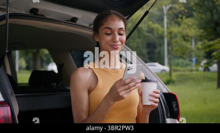 African American woman sitting trunk car auto automobile vehicle owner user mobile phone smartphone girl social media relaxing holding cup coffee Stock Photo