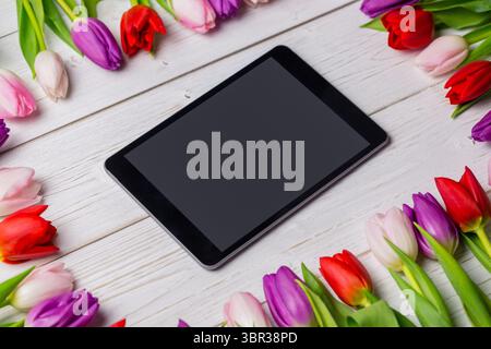 Showing flat lay featuring black tablet diagonally on white wooden tabletop framing tulip blooms Stock Photo
