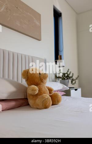 Teddy bear is sitting upright on bed with white sheets, pink pillows, abstract art, near nightstand Stock Photo