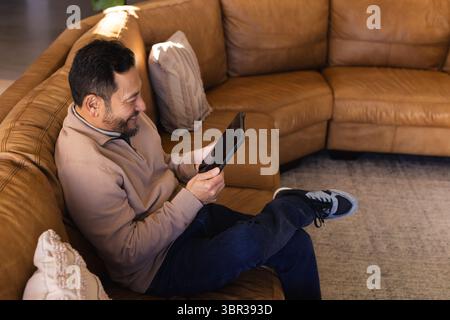 Asian man sitting on leather sectional sofa at home holding tablet with pillows wearing sneakers Stock Photo