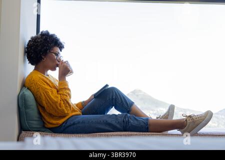 woman sitting on window seat with blue pillow sipping from mug and reading tablet mountain view Stock Photo