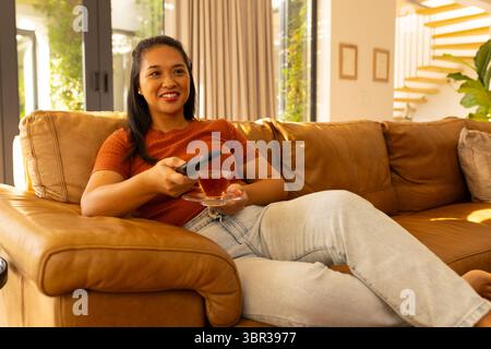 Asian woman reclining on sofa in living room, holding TV remote and glass teacup with saucer Stock Photo