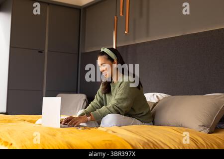 Asian woman sitting cross-legged on mustard yellow bedspread typing on silver laptop in bedroom Stock Photo