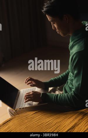 Man typing on slim laptop on mustard cushion on floor, side light casting shadows, curtains behind Stock Photo