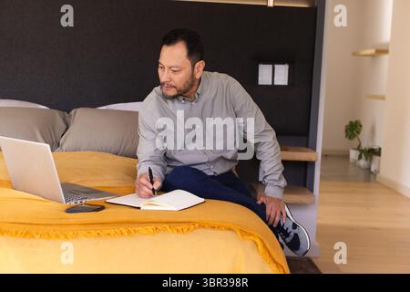 Asian man sitting on bed in bedroom writing in notebook near laptop and smartphone Stock Photo