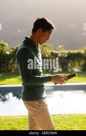 Asian man standing beside swimming pool in backyard wearing green sweater and using smartphone Stock Photo
