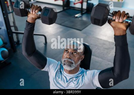 Senior African American man performing overhead dumbbell press on bench in gym with rubber floor Stock Photo