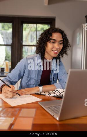Non-binary adult sketching designs on paper at home kitchen table, using laptop and color swatches Stock Photo