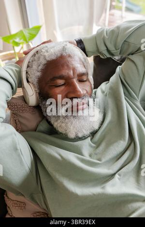 Senior African American man reclining on sofa at home, using headphones and checking smartwatch Stock Photo