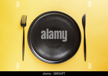 Black ceramic plate with knife and fork on yellow background. Pop art design Stock Photo