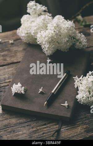 Spring romantic composition with white lilac flowers and old notebook for sketches on dark wooden background Stock Photo