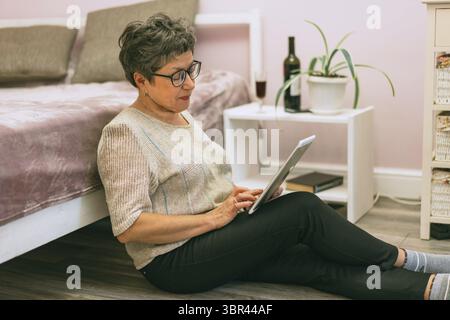 Attractive elegant senior woman sitting on the floor near the bed and using a tablet computer at home Stock Photo