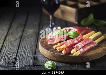 Bread sticks grissini with prosciutto ham on cutting board over wooden background, close up Stock Photo