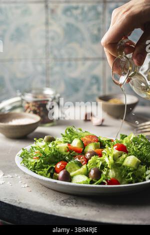 A hand pouring olive oil into classic vegetable salad with fresh olives, tomatoes and cucumbers on gray background Stock Photo