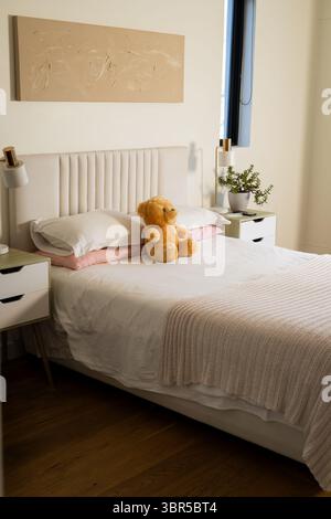 Plush golden brown teddy bear sitting on upholstered bed with knitted throw at bedroom window Stock Photo