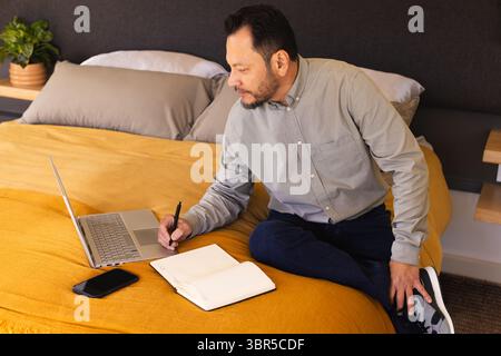 Mature Asian man sitting on bed using laptop and writing in notebook with smartphone nearby Stock Photo