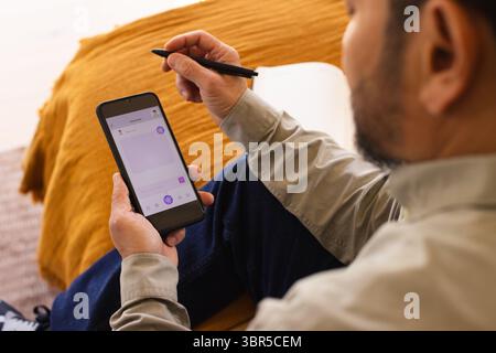 Middle-aged Asian man sitting on sofa with orange throw sending message using smartphone stylus pen Stock Photo
