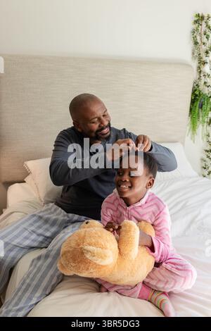 African American father wearing pajamas sitting on bed braiding daughter's hair holding teddy bear Stock Photo