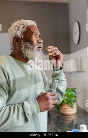Senior African American man holding pill and glass of water by home kitchen countertop Stock Photo