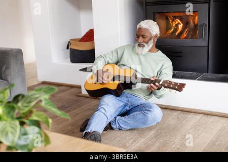 Senior African American man wearing green sweatshirt strumming acoustic guitar at home by fireplace Stock Photo