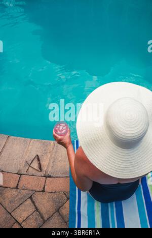 Woman in hat and swimsuit sitting on poolside with drink in summer. Stock Photo