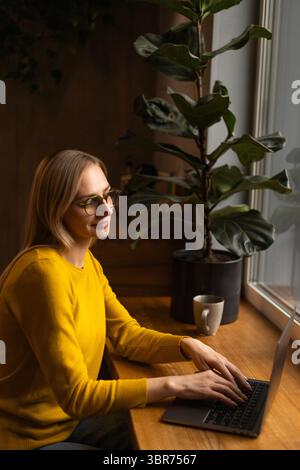 A woman in a mustard sweatshirt is smiling while working on a laptop. Stock Photo