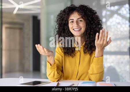 A cheerful woman with curly hair, gestures expressively while smiling, possibly presenting or explaining something in an office setting. Stock Photo
