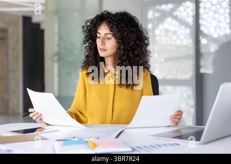 A woman with curly hair examines documents at her desk, appearing focused on her work in a bright office setting. Stock Photo