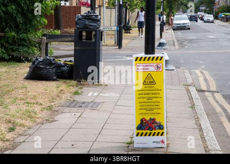 Wide shot of a yellow "No Dumping Rubbish" sign on a lamp post, with dumped rubbish scattered around a public waste bin behind—highlighting urban litt Stock Photo