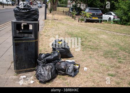 An overflowing rubbish bin surrounded by scattered bags of trash on the ground highlighting urban waste Stock Photo