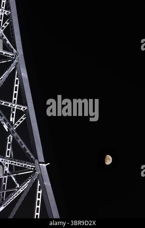 Juxtaposed against glowing waxing gibbous moon, steel beams of a television tower stretch vertically upward into the darkness of a starless night sky. Stock Photo