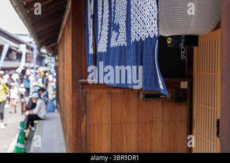 Japanese Noren curtain hangs in doorway of traditional restaurant with aged wooden exterior. Festival goers line streetside in front of building. Stock Photo