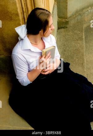 A woman in a white shirt and black skirt is sitting on some steps, holding a book. Stock Photo