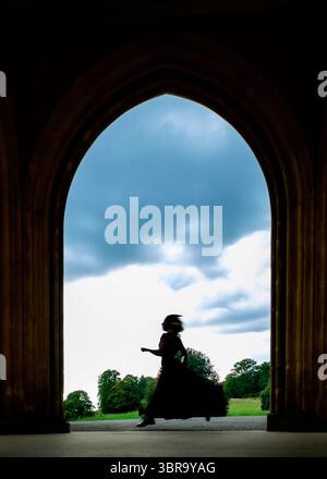 A silhouetted figure in a flowing dress runs through an archway towards a green field and cloudy sky. Stock Photo