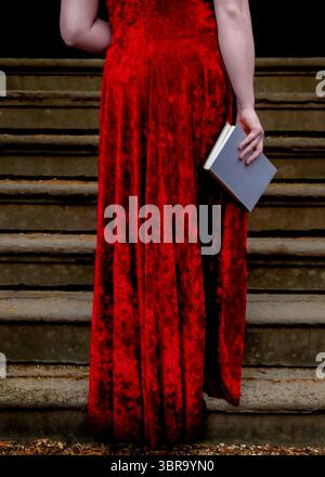A person in a long red dress stands on a set of stairs, holding a book. Stock Photo