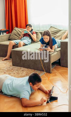 blurred boy lying on wooden floor playing with tablet while two girls relax on sofa using smartphones with orange curtains and daylight, technology Stock Photo