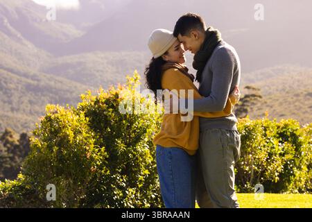 Couple embracing on grassy hillside wearing knit sweaters and scarves overlooking misty mountains Stock Photo