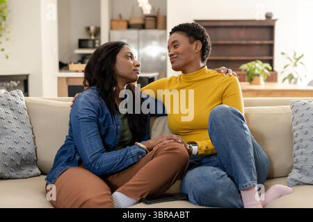 Diverse female friends sitting on beige sofa at home scrolling smartphone among textured cushions Stock Photo