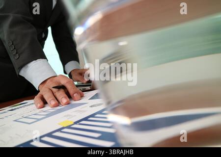 Close-up of a businessman in a suit leaning over a desk while analyzing printed financial graphs and data charts. A pen rests in his hand as he review Stock Photo