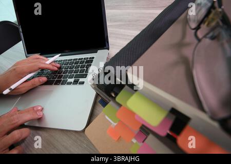 A close-up top view of a person working on a laptop with one hand holding a digital stylus. A tall stack of folders and notebooks with colorful index Stock Photo