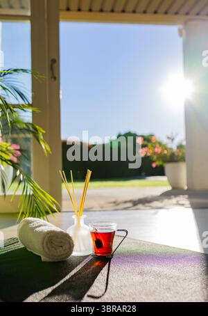 Yoga mat lying on covered porch during morning light, holding rolled towel, reed diffuser, tea mug Stock Photo