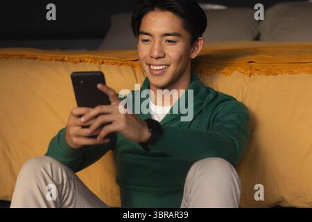 Asian man sitting on living room floor leaning on mustard yellow sofa smiling holding smartphone Stock Photo