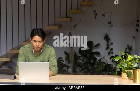 Asian man typing on silver laptop at counter with tablet, philodendron, brass container, terrarium Stock Photo