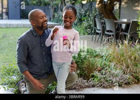 Black father kneeling beside daughter blowing bubbles from pink container on patio Stock Photo