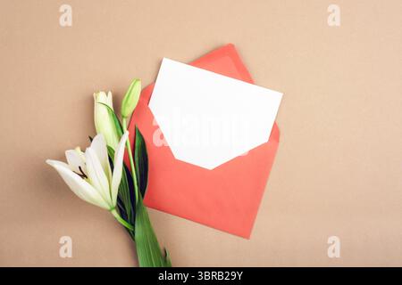 White blank card in red envelope with white lily flowers on beige background. Top view, flat lay, mockup. Stock Photo